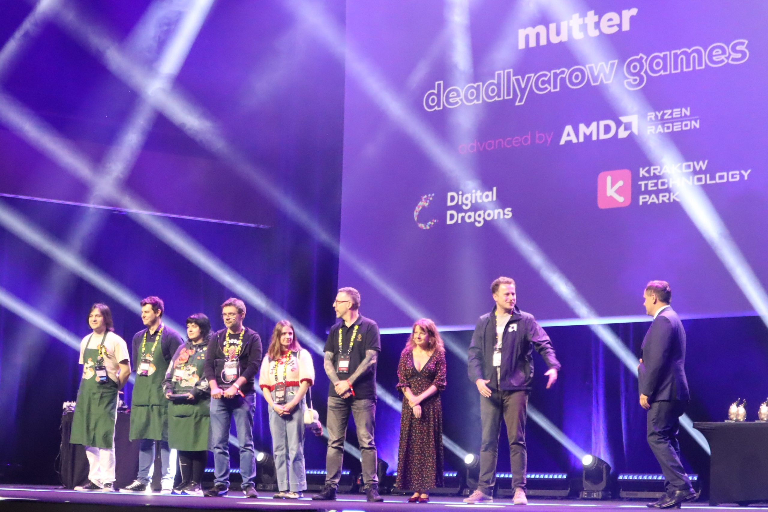 Group of people standing on a stage under purple lighting, event backdrop shows sponsor logos like Digital Dragons and AMD/Ryzen Radeon.",
