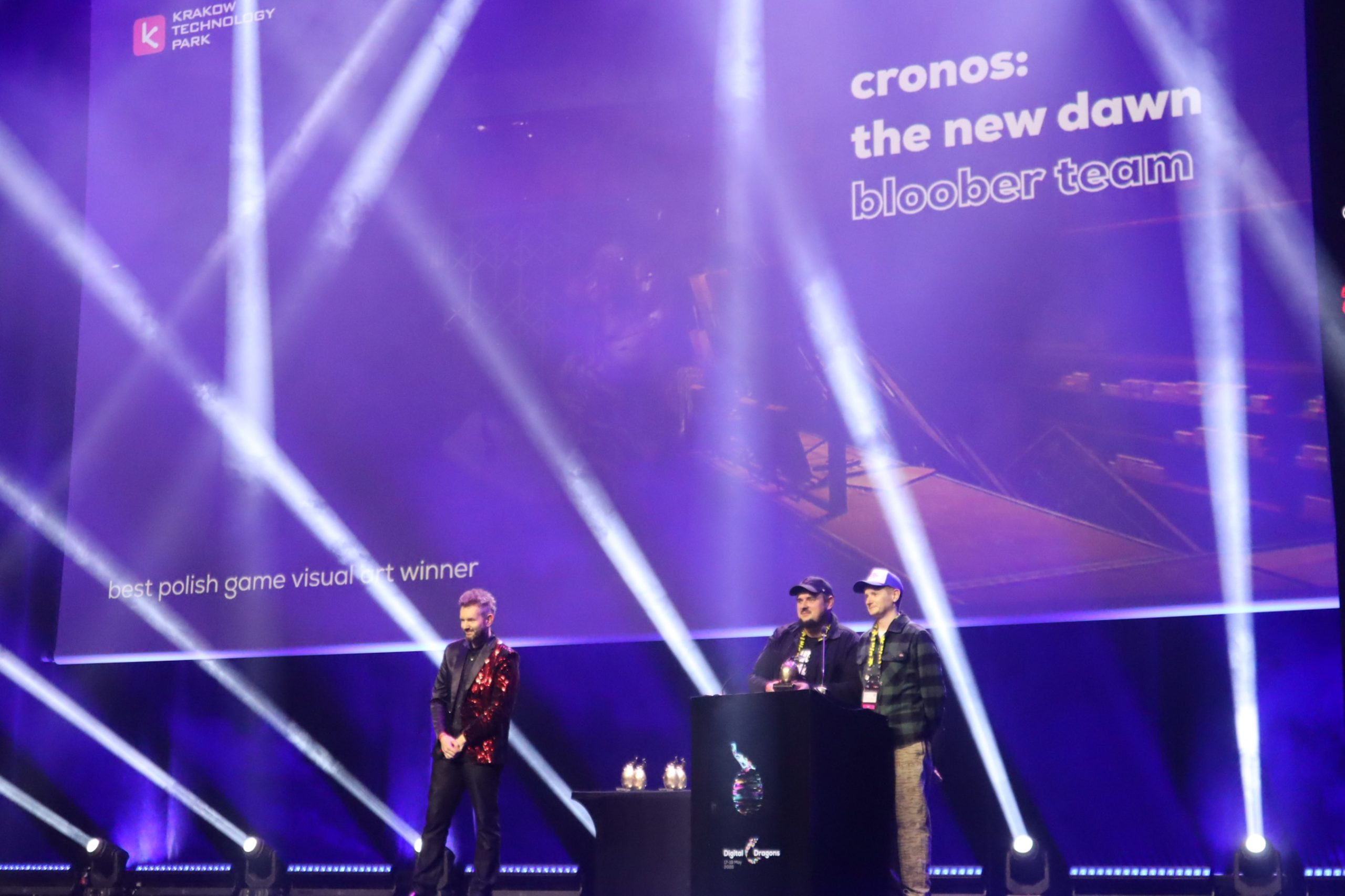Stage at an awards ceremony with purple lighting; text on screen reads 'cronos: the new dawn bloober team' and logos in top left, three men standing on stage with a podium.