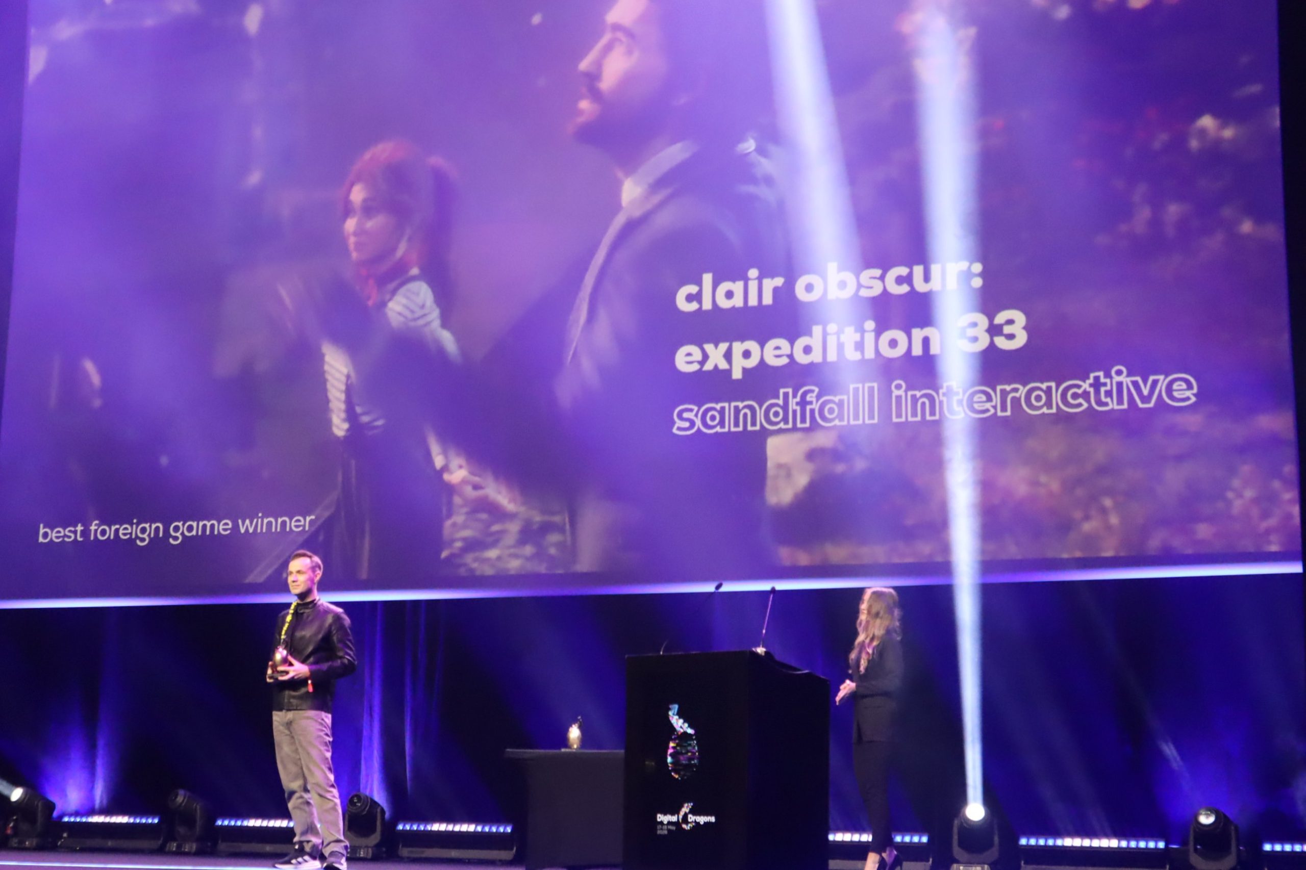 Stage with two presenters under purple lighting, backdrop shows 'clair obscur: expedition 33 sandfall interactive'