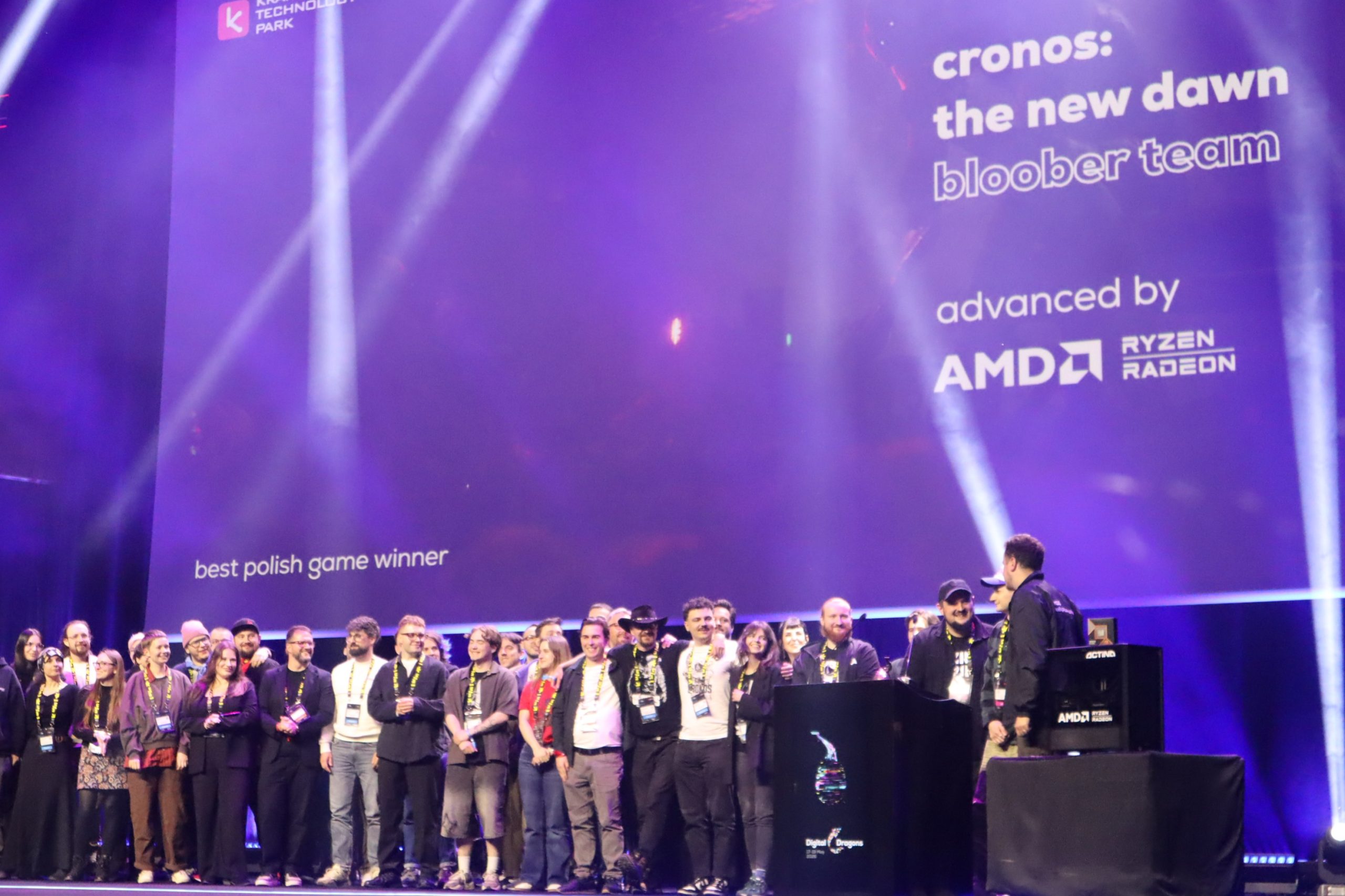 Large group of people posing on a stage at a tech event with purple backdrop and sponsor logos.