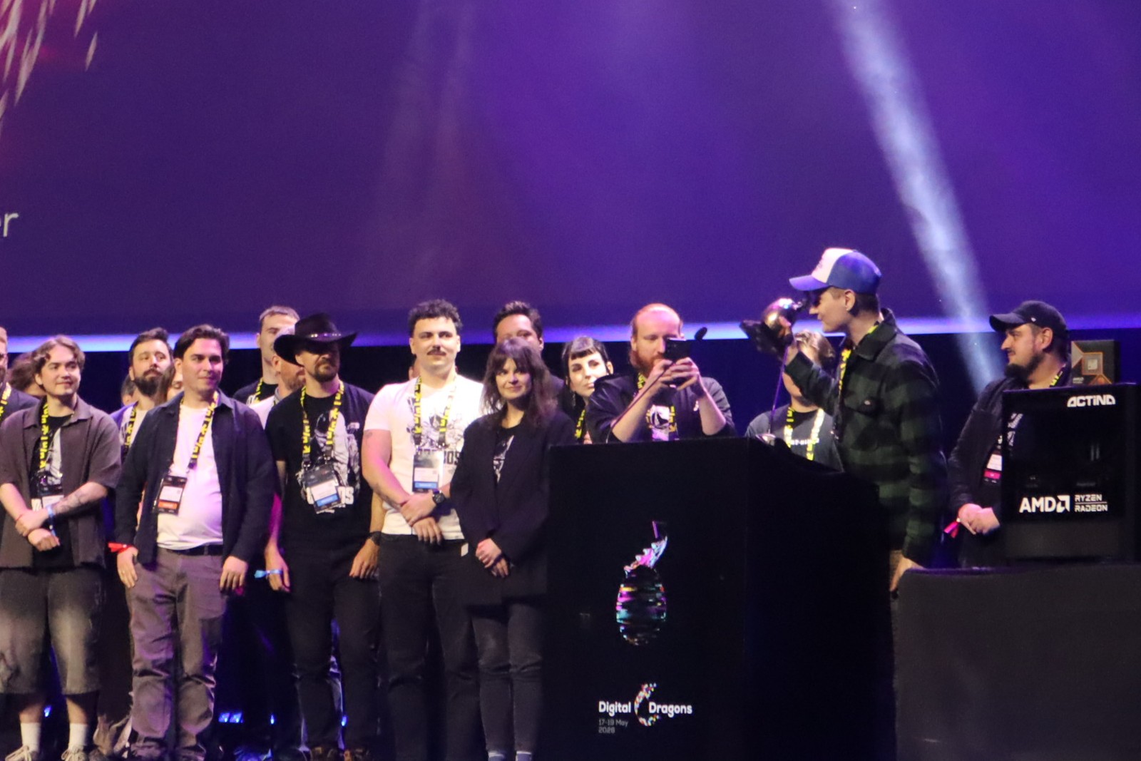 Group of people on a stage at a tech event, some wearing lanyards, with a speaker at the podium and others watching.