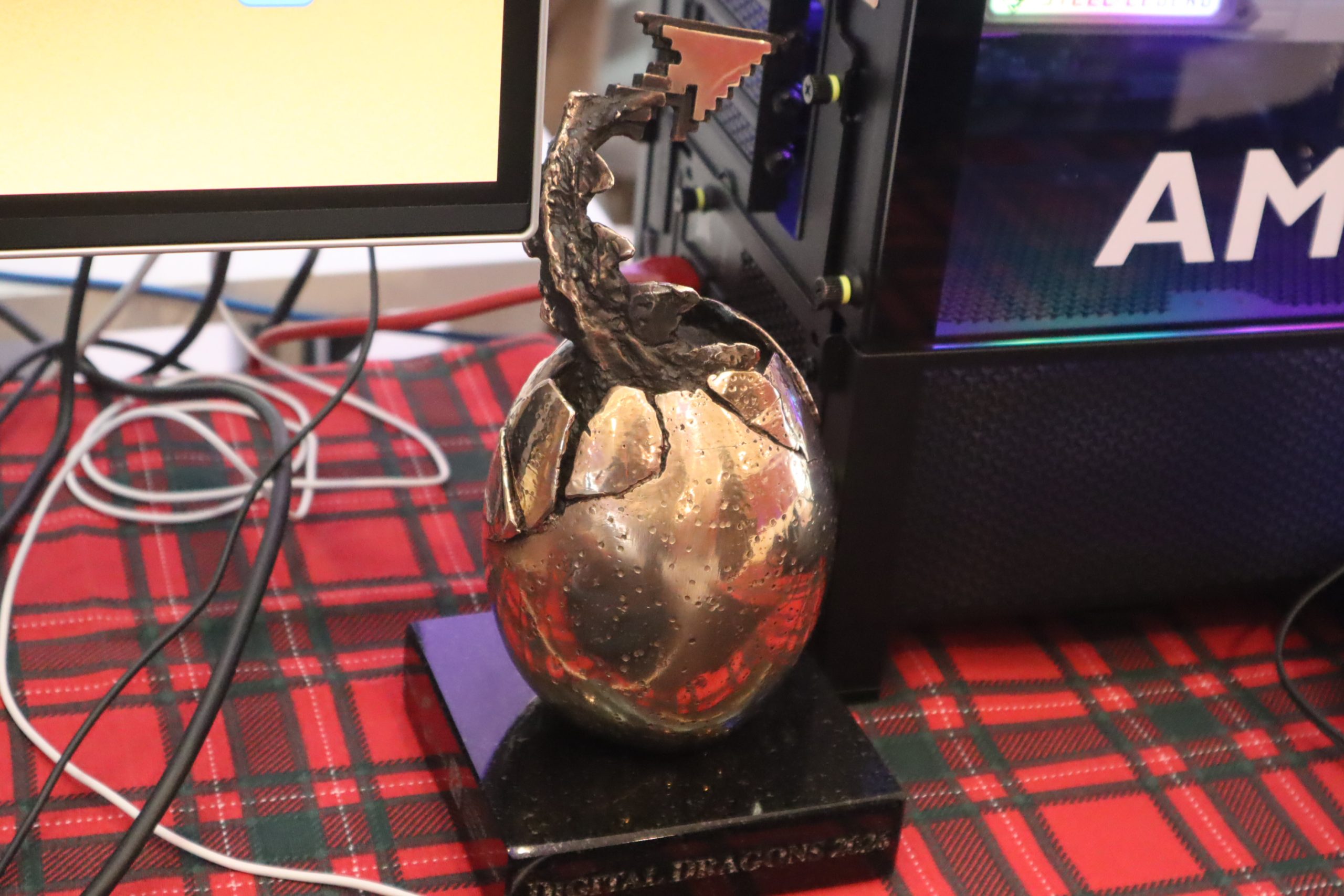Broken bronze trophy with a jagged top, on a black pedestal, resting on a red plaid table near computer cables and equipment.