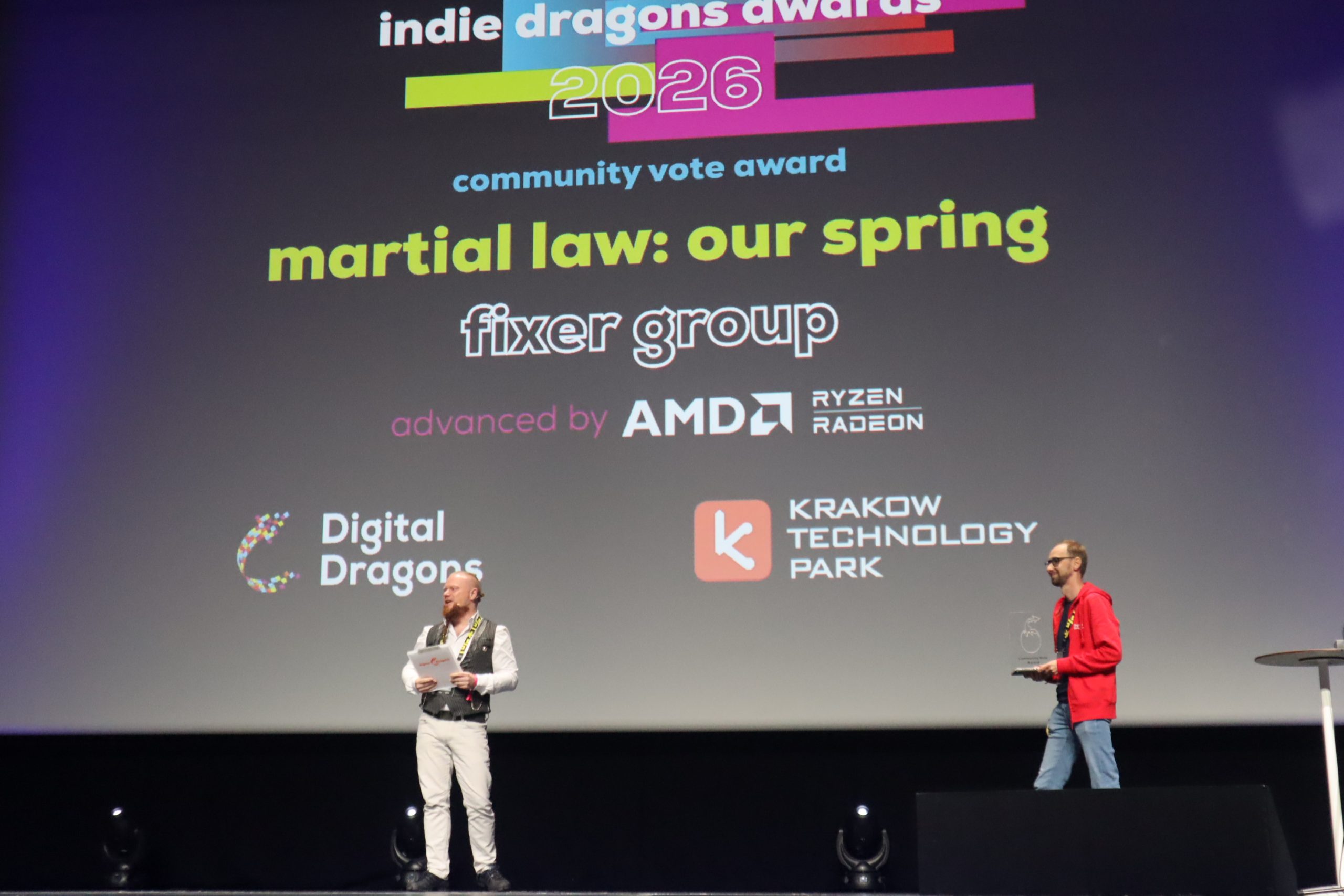 Two men stand on a stage presenting an award, with a large colorful screen showing logos and text behind them.