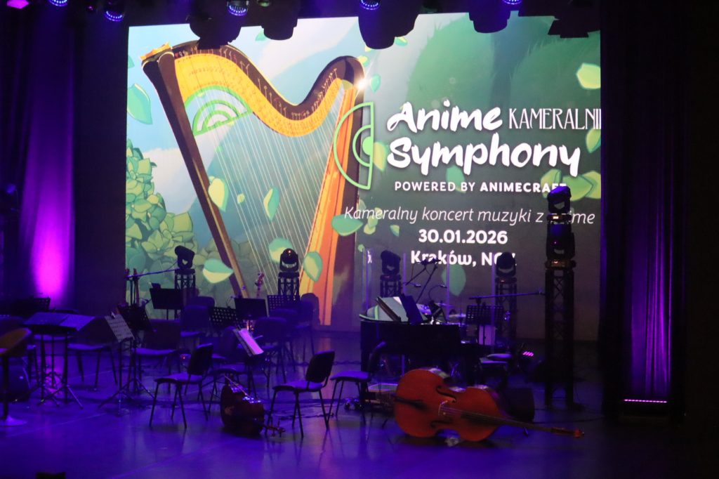Stage for an Anime Symphony concert, with a large colorful screen showing a harp illustration and the words 'Anime Symphony' amid green foliage, and rows of music stands on stage under purple lighting.