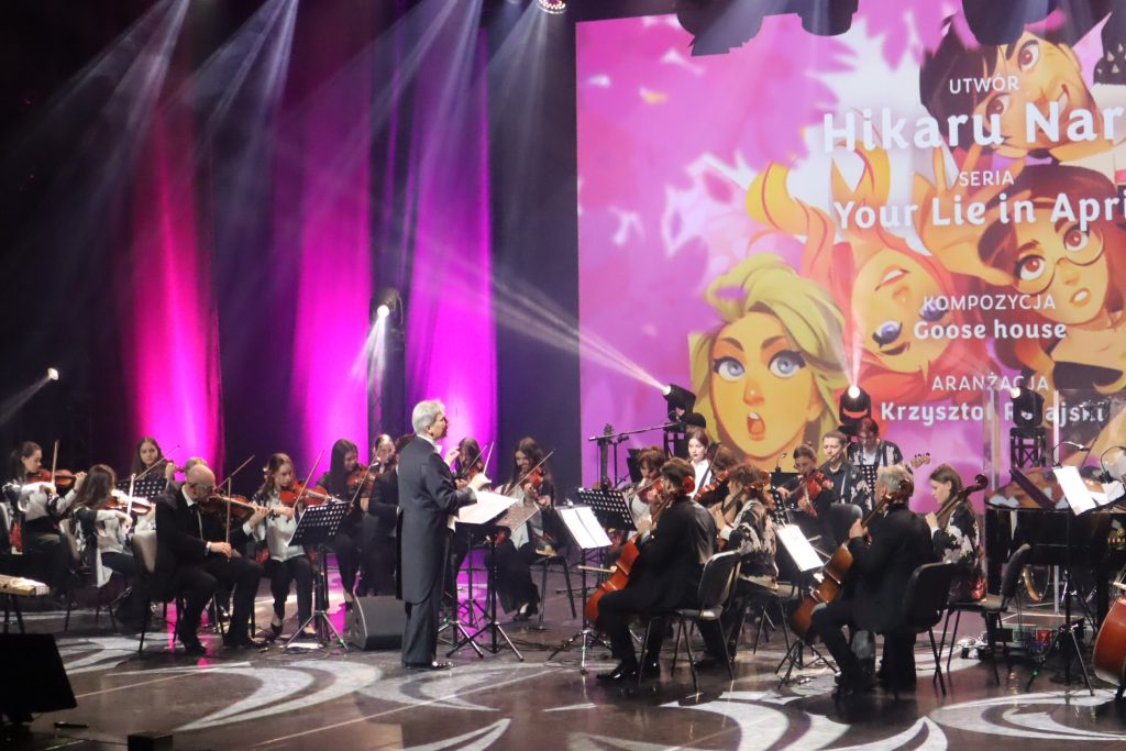 Conductor leads a large orchestra on stage with purple lighting and a colorful backdrop behind them.