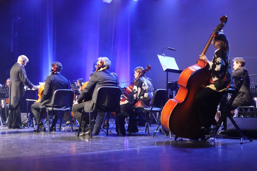 Orchestra performing on stage under blue lights, conductor at left and string players with cellos and basses visible.