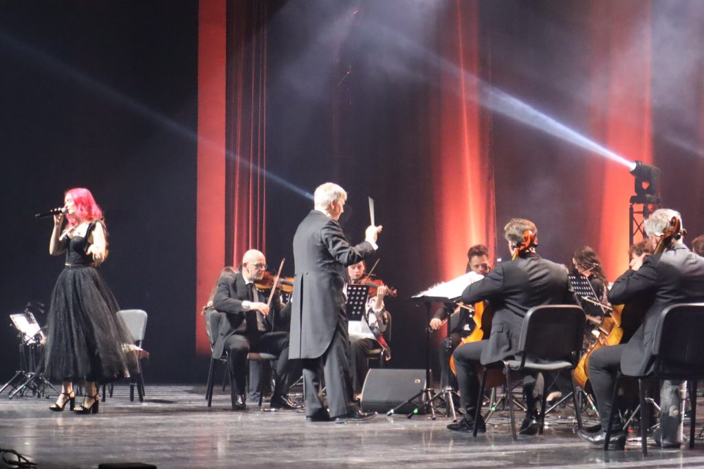 Female singer with pink hair performs on stage beside an orchestra, conductor directing amid red stage lighting, instruments visible on a dark backdrop.
