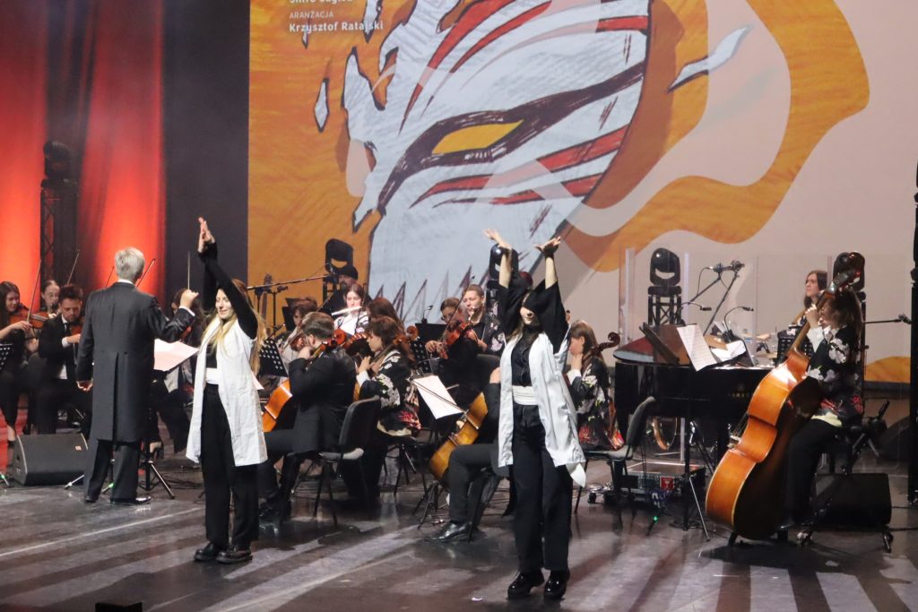 Conductor leading a large orchestra on stage, musicians playing string instruments under warm orange stage lighting with a large abstract backdrop behind them.