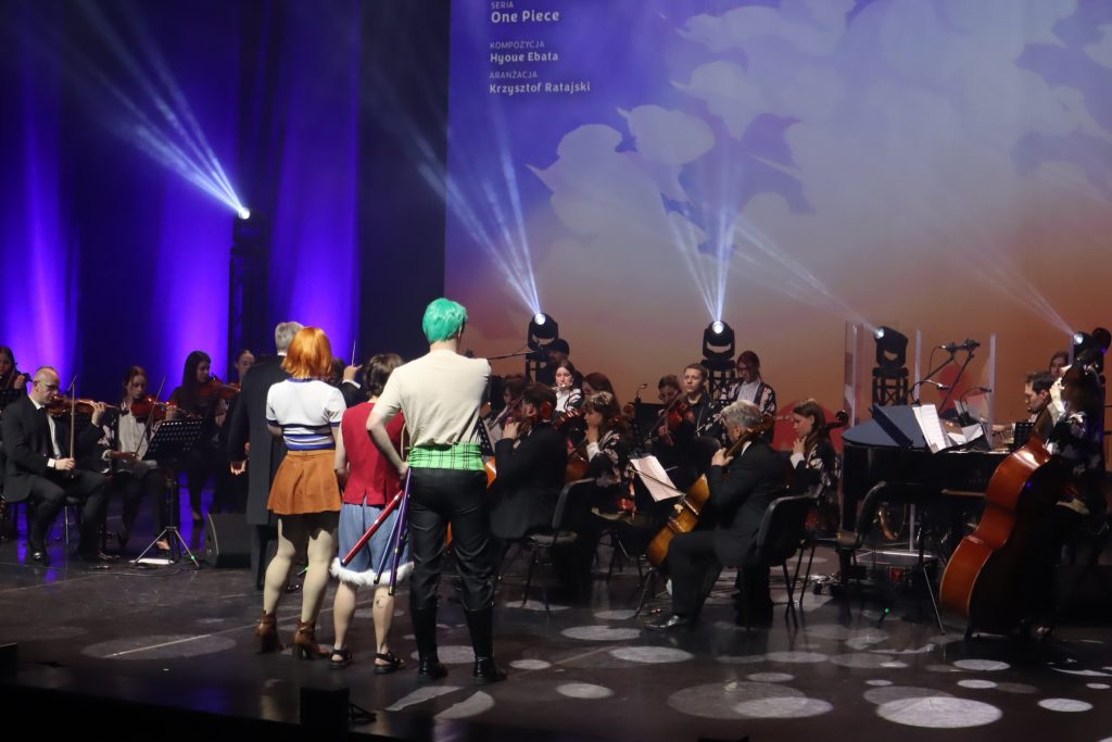 Orchestra performing on a stage with a conductor and musicians in colorful outfits, purple stage lights and a large screen in the background.