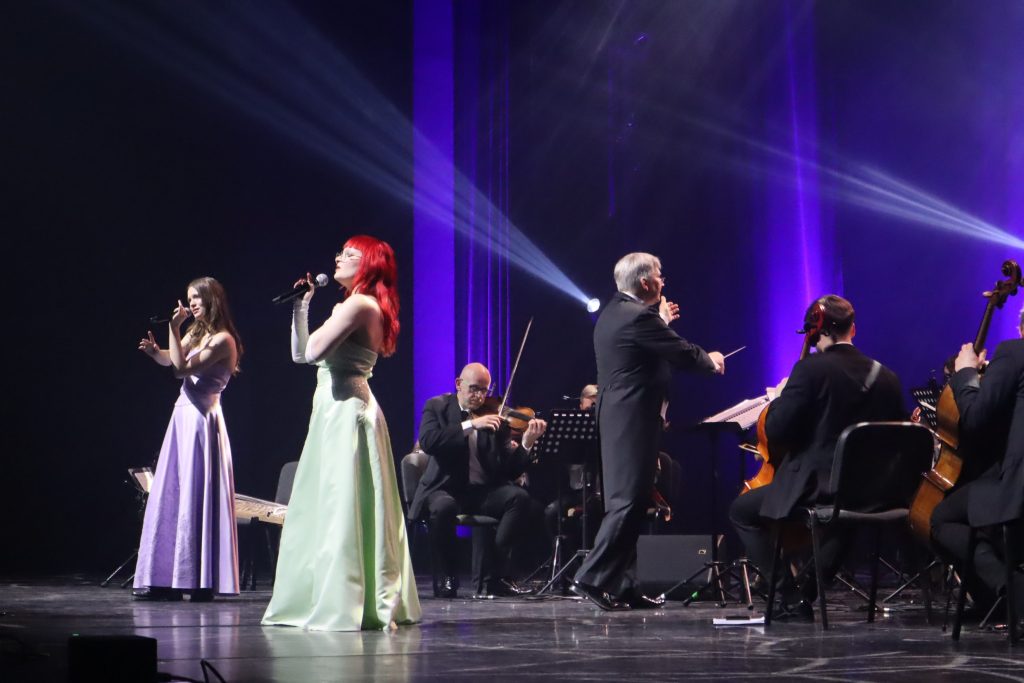Two female singers in long pastel gowns perform center stage with microphones, backed by a full orchestra on a dark stage. Purple lighting washes the background.