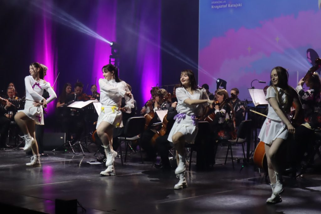 Dancers in white outfits perform a choreographed routine on stage as an orchestra plays behind them, bathed in purple lighting.
