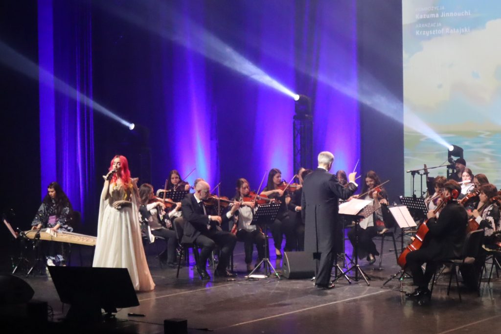 Female singer in a white gown performs with a large orchestra on a stage bathed in blue lighting.
