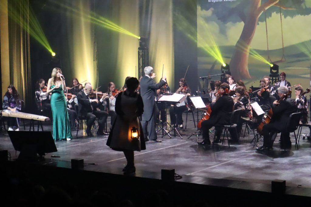 Conductor leads an orchestra on stage as a female singer in a green gown performs beside violinists under yellow stage lights.