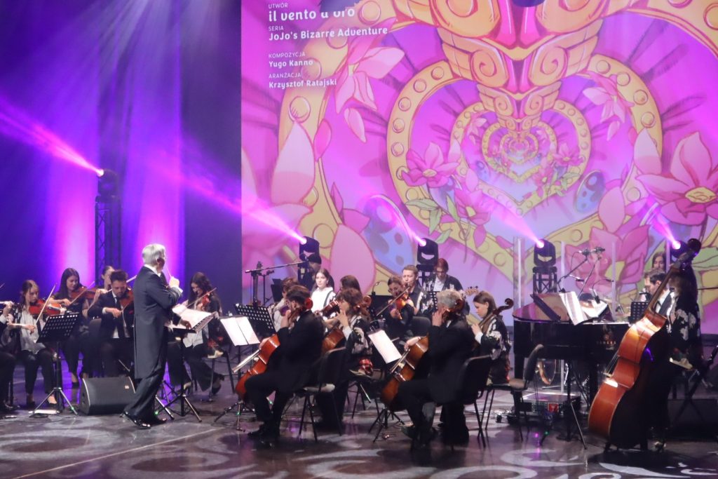 Conductor leads a large orchestra on a dimly lit stage with purple spotlights and a vibrant mural backdrop.