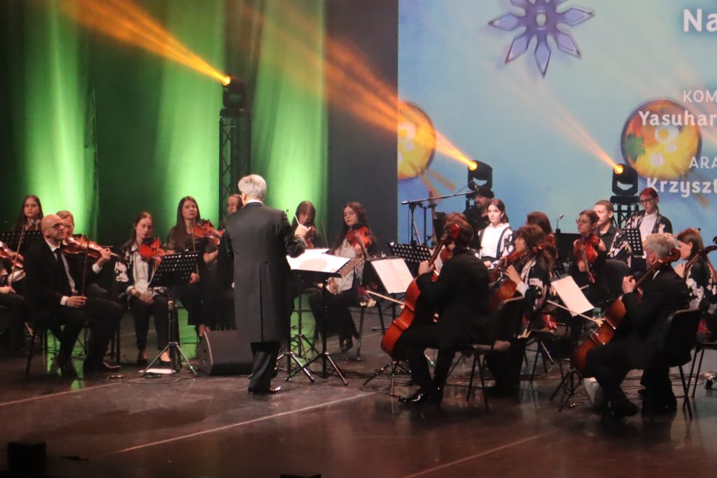 Orchestra on stage led by a conductor, string players with violins and cellos under green lighting and a blue backdrop.