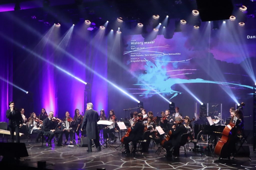 Orchestra performing on a dimly lit stage with purple and blue lights; conductor in front guiding musicians.