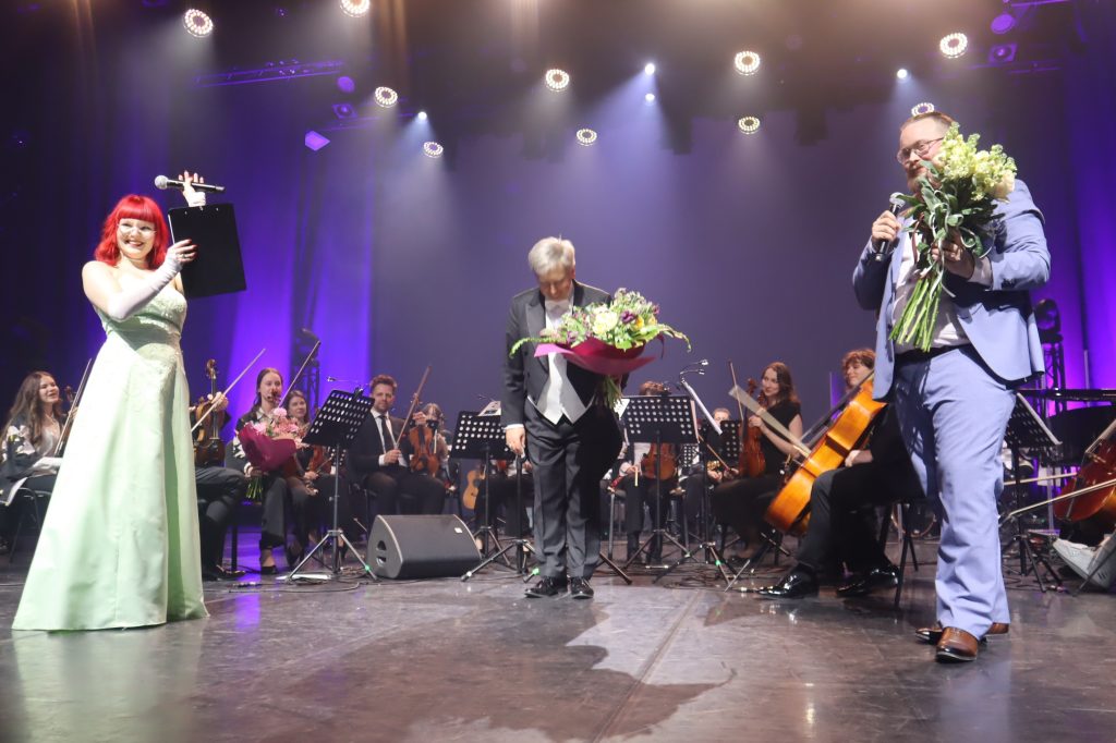 Female singer in a pale green gown holds a microphone on stage with an orchestra behind her, bright spotlights above.
