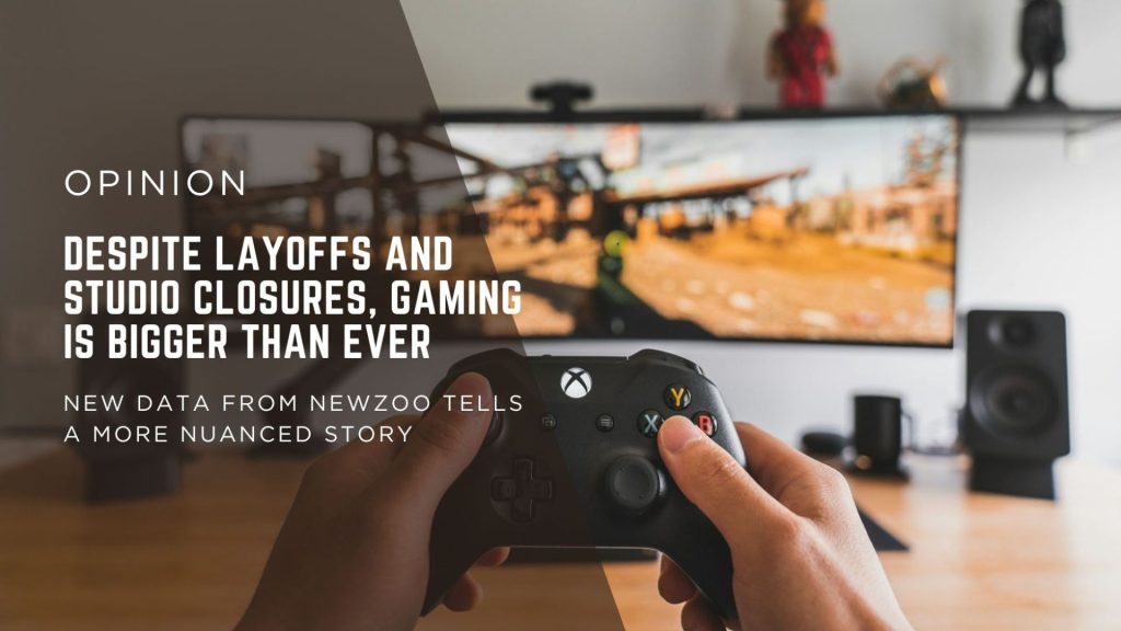 Person's hands grip a black game controller in the foreground with a blurred desk setup and a large monitor, matching a headline on layoffs and gaming (opinion).