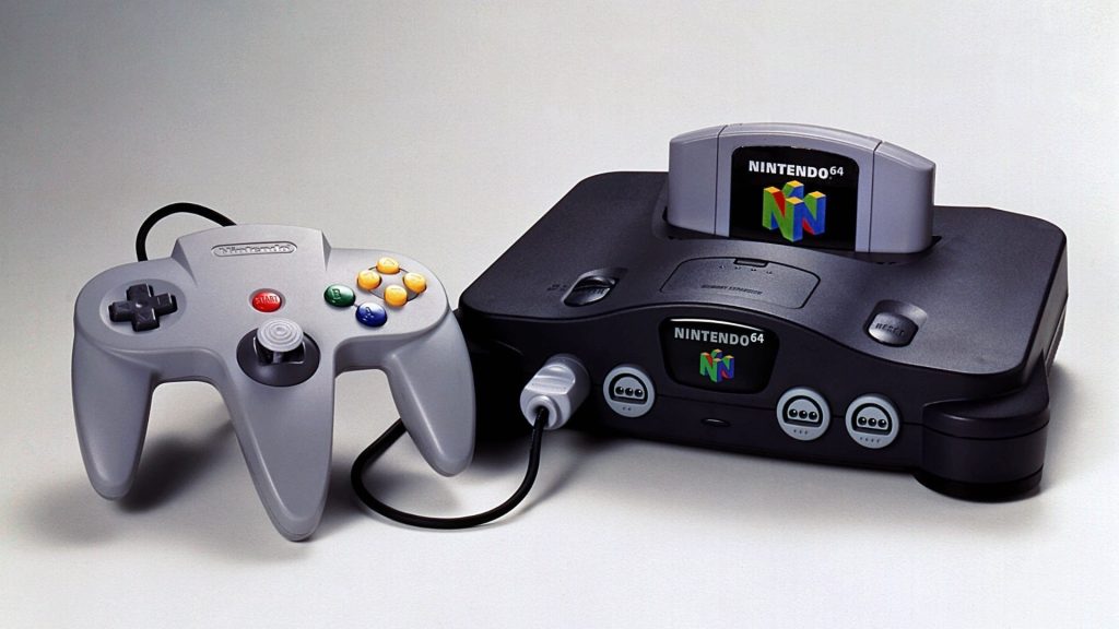 Nintendo 64 game console with a wired controller in front and a cartridge inserted in the top slot on a light background.