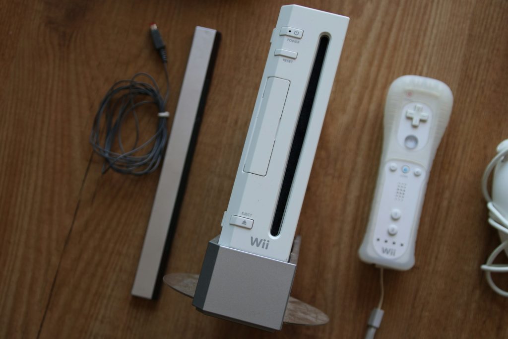 White Nintendo Wii console upright on a stand with a remote and cables on a wooden floor, including a sensor strip nearby.